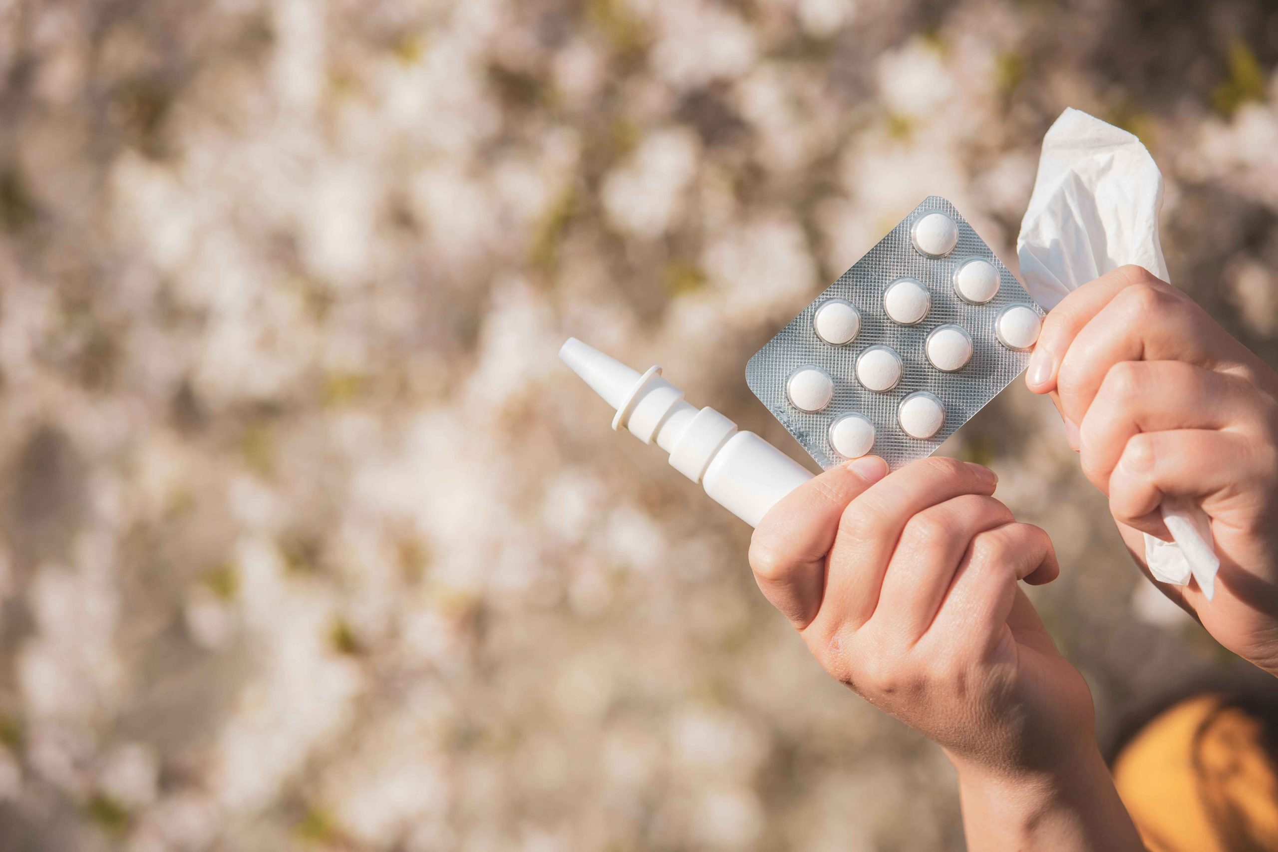 Hands holding allergy medications, including a nasal spray, blister pack of tablets, and a tissue, set against a blurred outdoor background, indicating allergy relief and treatment.